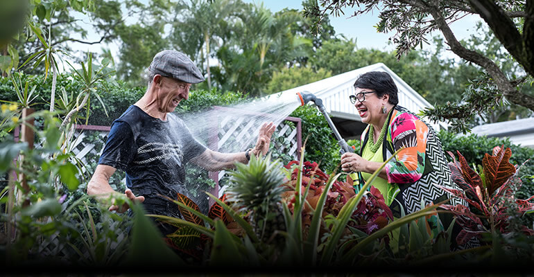 Woman in brightly patterned top playfully hosing man wearing a grey hat in the garden.