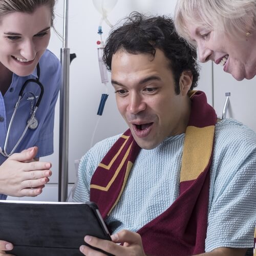 A surprised man in hospital gown and sports scarf with two nurses looking down at an iPad watching sports