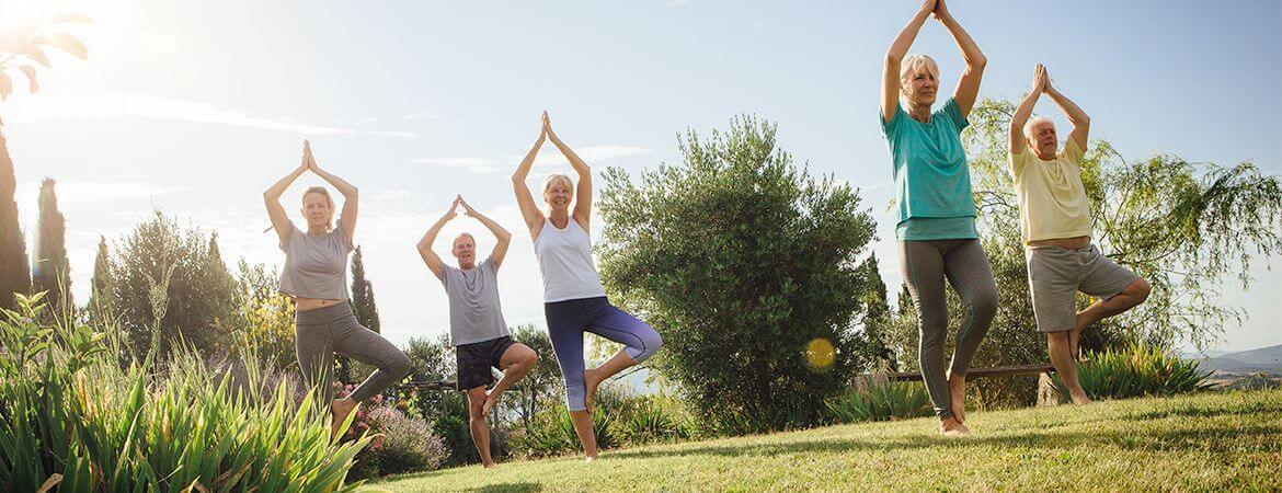 Two men and three women in a park, holding up their hands and standing on their right leg in a yoga pose.