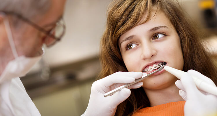 A close-up of an elderly male dentist treating the mouth of a young girl with braces wearing an orange shirt in a dentist surgery. 