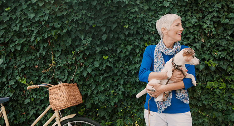 Elderly woman stands next to bike, holding dog in hands and looking off to her left, with a leafy background.