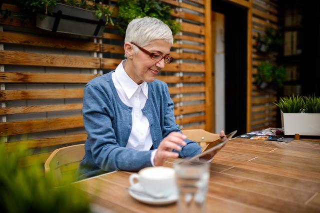 Mature woman sitting outside cafe drinking coffee and using digital tablet