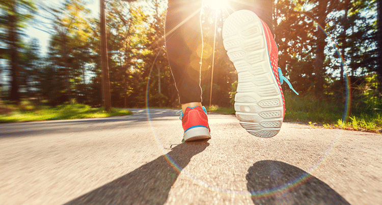 A close-up of a person mid-stride running on the road wearing red and blue runnings shoes and green trees in the background. 