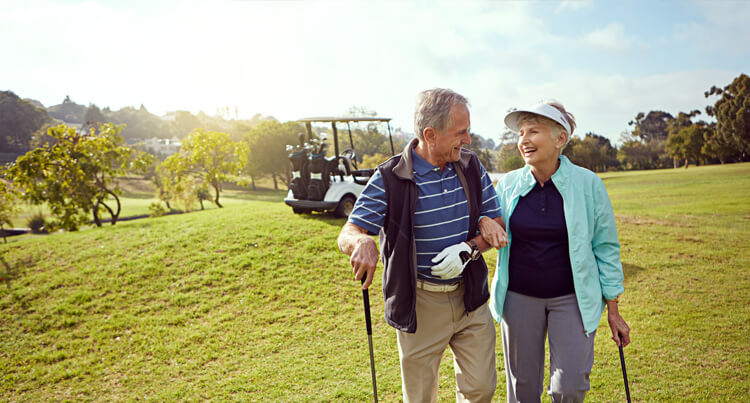 An elderly man and woman linking arms on a golf course smiling in front of a golf buggy, with green grass and trees in the background