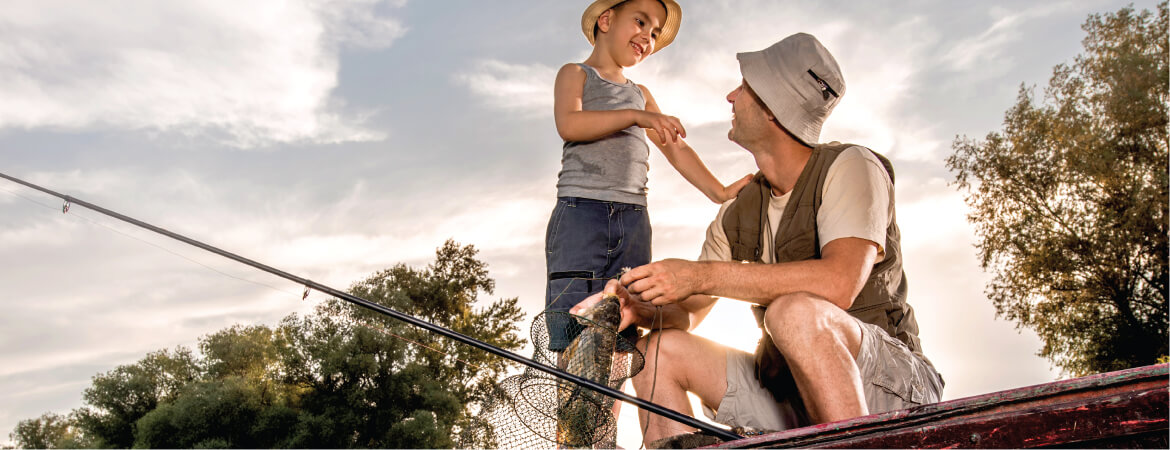 Small boy and man in a vest wearing hats looking at each other and holding a fishing rod and net while sitting in a boat